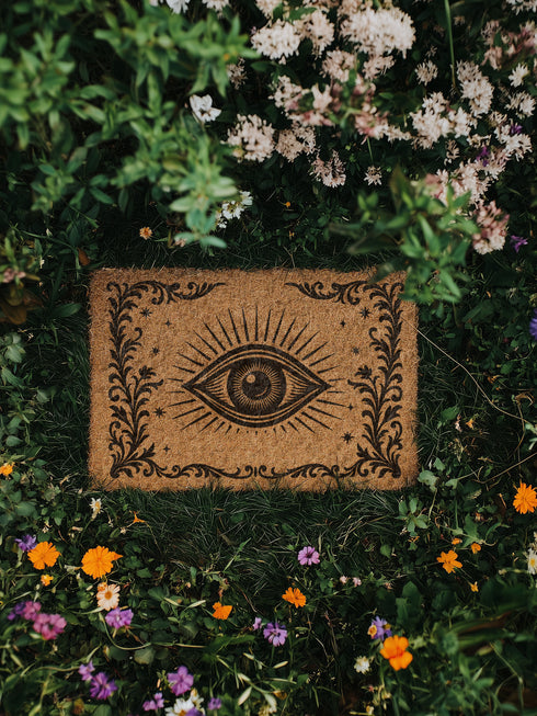 A 24x16 inch rectangular outdoor doormat made of eco-friendly coconut coir with a black vinyl backing. The mat features a central evil eye design radiating with detailed rays and surrounded by swirling ornamental scrollwork and small stars. Designed for witchy home entryways, it offers protection and style for porches, patios, and thresholds.